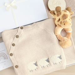 A beige knitted baby cardigan with brown buttons and embroidered sheep, knitted beige footed trousers with a giraffe plush toy, a gift box, and a wooden age milestone plaques.